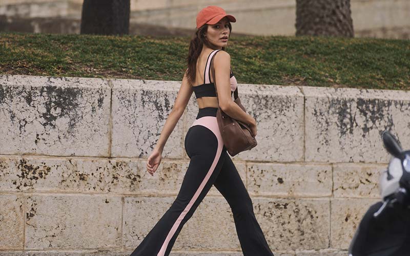 Woman in black and pink activewear walking outdoors with a red cap and crossbody bag.