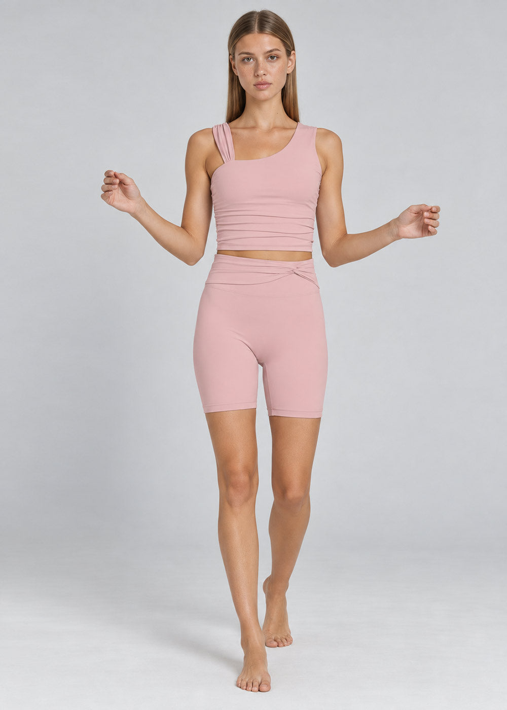 Woman wearing a soft pink activewear set with an asymmetrical strappy tank top and high-waisted fitted biker shorts, shown from the front in a full-body walking pose.