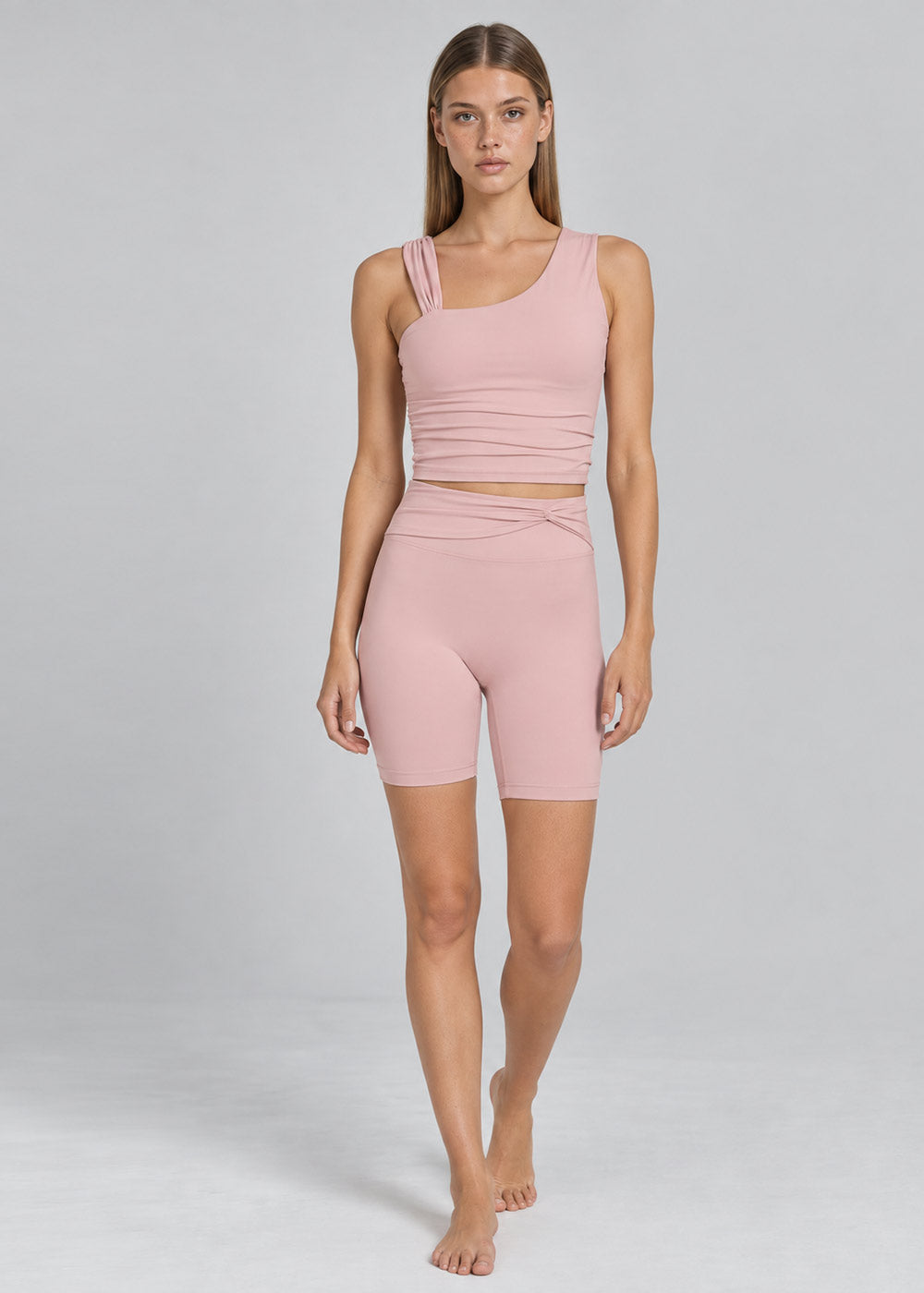 Woman wearing a soft pink activewear set with an asymmetrical strappy tank top and high-waisted fitted biker shorts, shown from the front in a full-body walking pose.