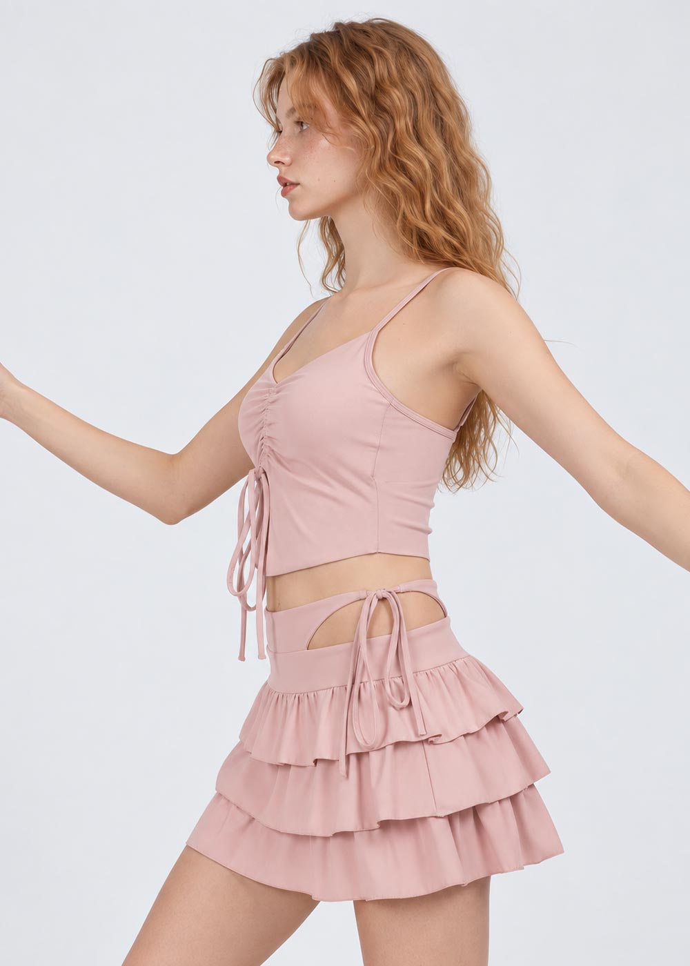 Side view of women's blush pink activewear set with ruched drawstring camisole top, layered ruffle skirt and waist cutout details