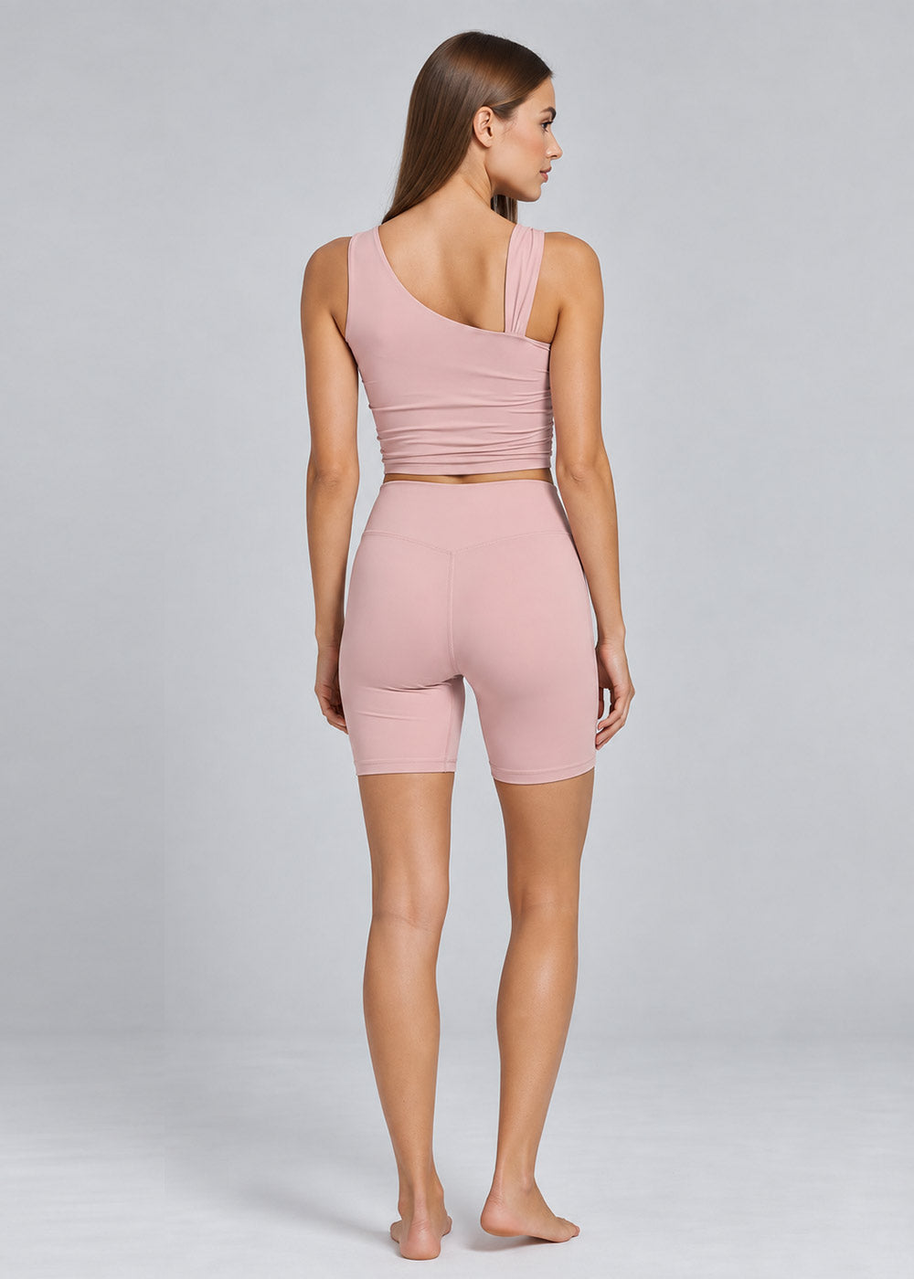 Woman wearing a soft pink activewear set with an asymmetrical strappy tank top and high-waisted fitted biker shorts, shown from the back in a full-body pose.