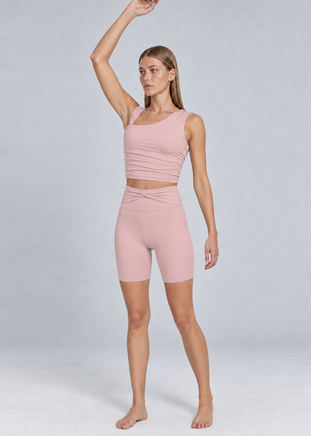 Woman wearing a soft pink activewear set with an asymmetrical strappy tank top and high-waisted fitted biker shorts, shown from the front in a full-body pose with one arm raised overhead.