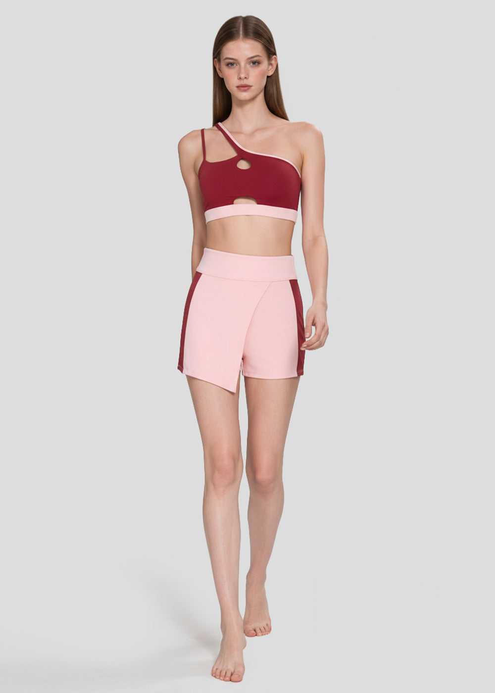 Women's burgundy one-shoulder sports bra with cutout details paired with blush pink high-waisted active shorts with contrast side panels