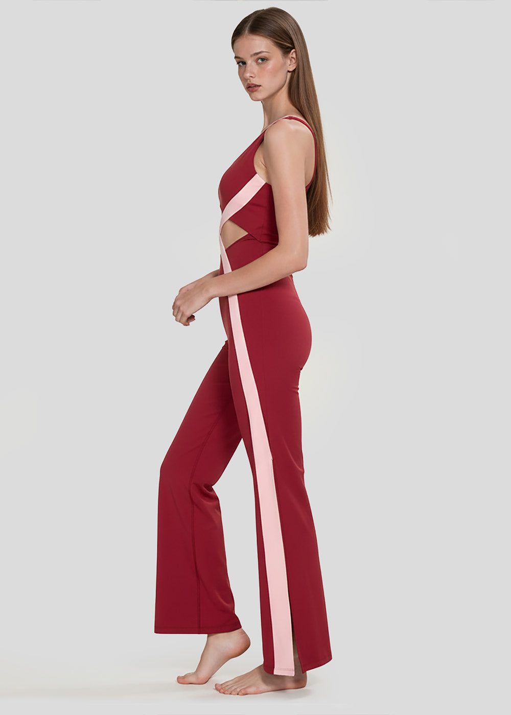 Side view of a model wearing a burgundy and blush pink women's wide-leg activewear jumpsuit with waist cutouts and contrast side stripes