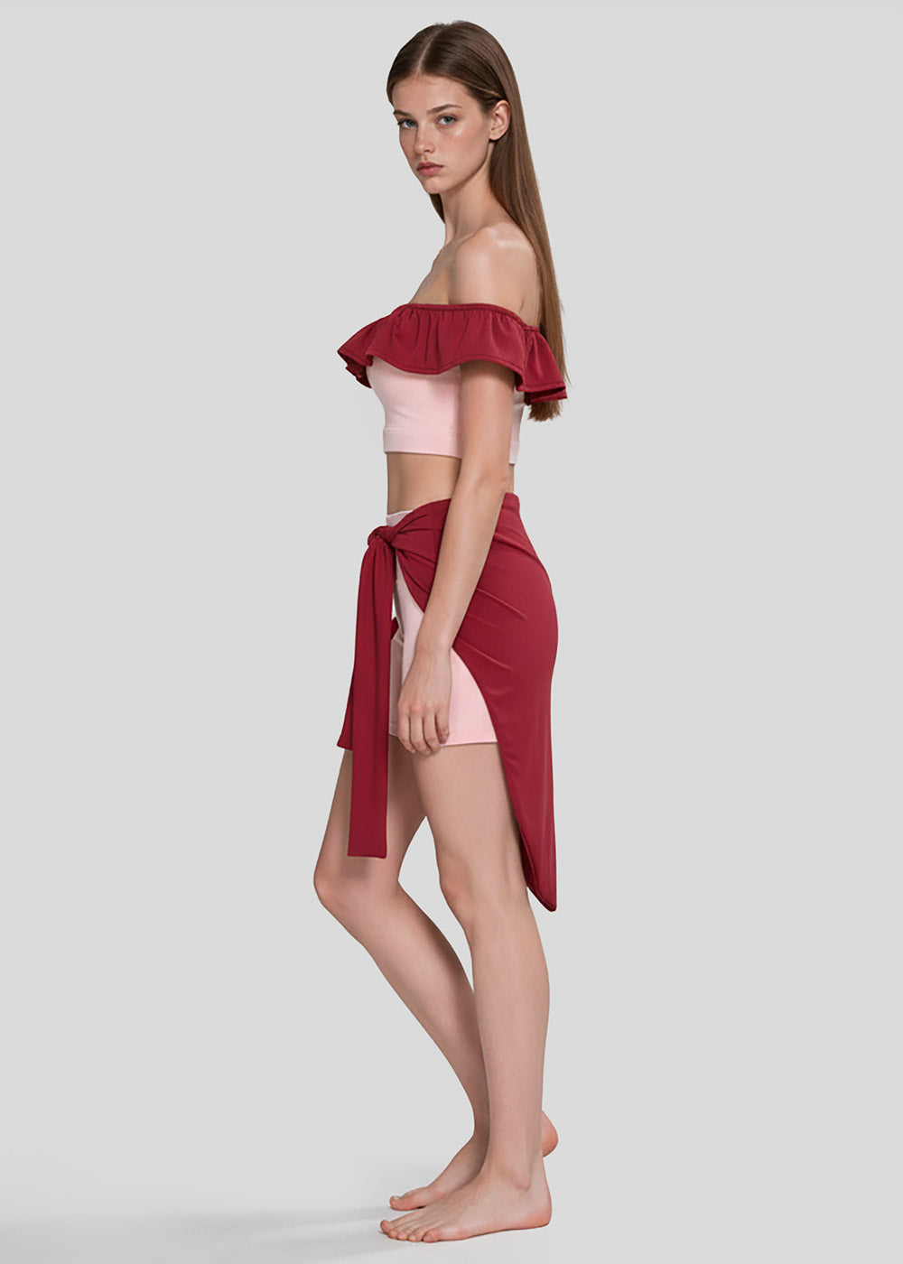Side view of women's blush pink off-shoulder ruffle crop top with burgundy trim, matching shorts and burgundy wrap skirt