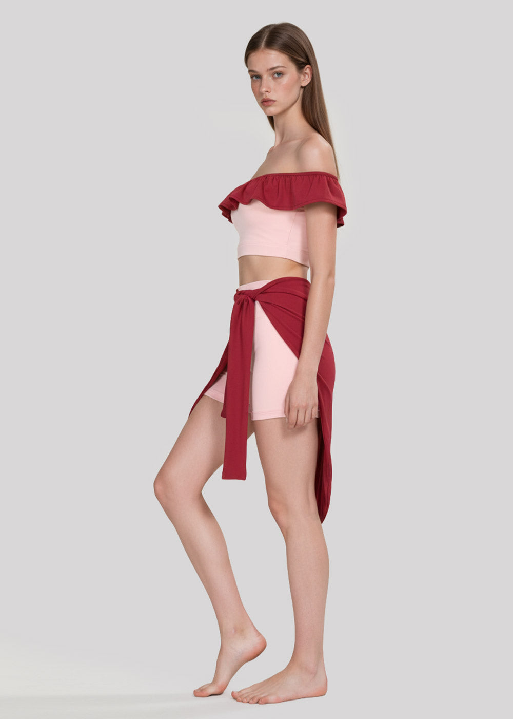 Side view of women's blush pink off-shoulder ruffle crop top with burgundy wrap skirt and fitted shorts