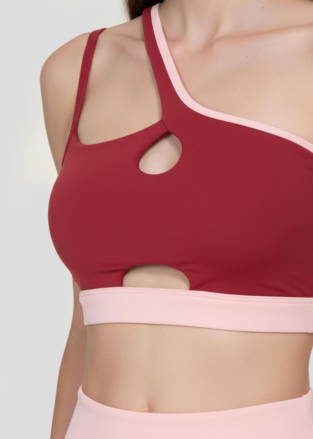 Close-up of a burgundy asymmetrical sports bra for women featuring double straps, cutout details and blush pink contrast trim
