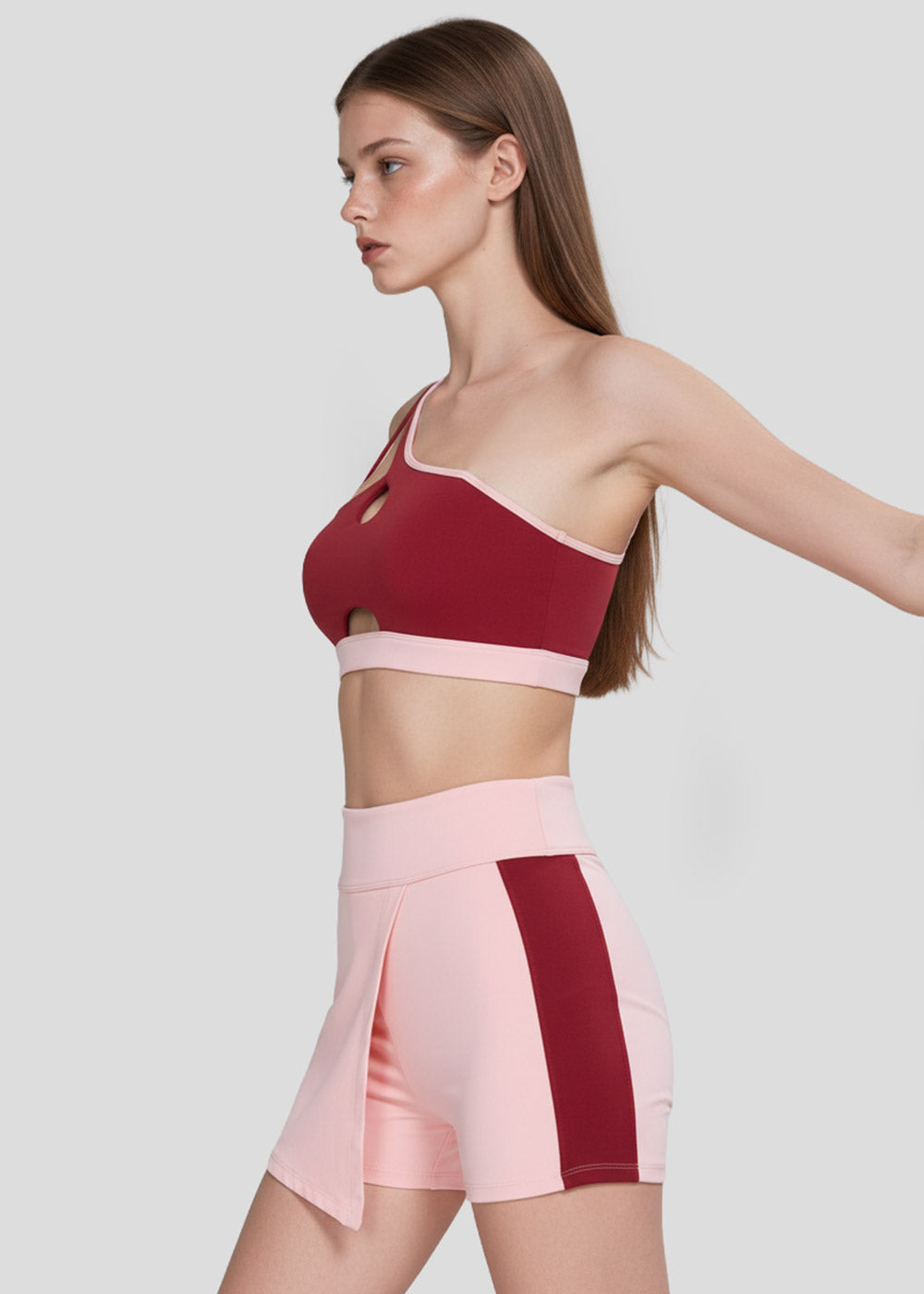 Women's burgundy asymmetrical sports bra with cutout details and blush pink high-waisted workout shorts with contrast side panels
