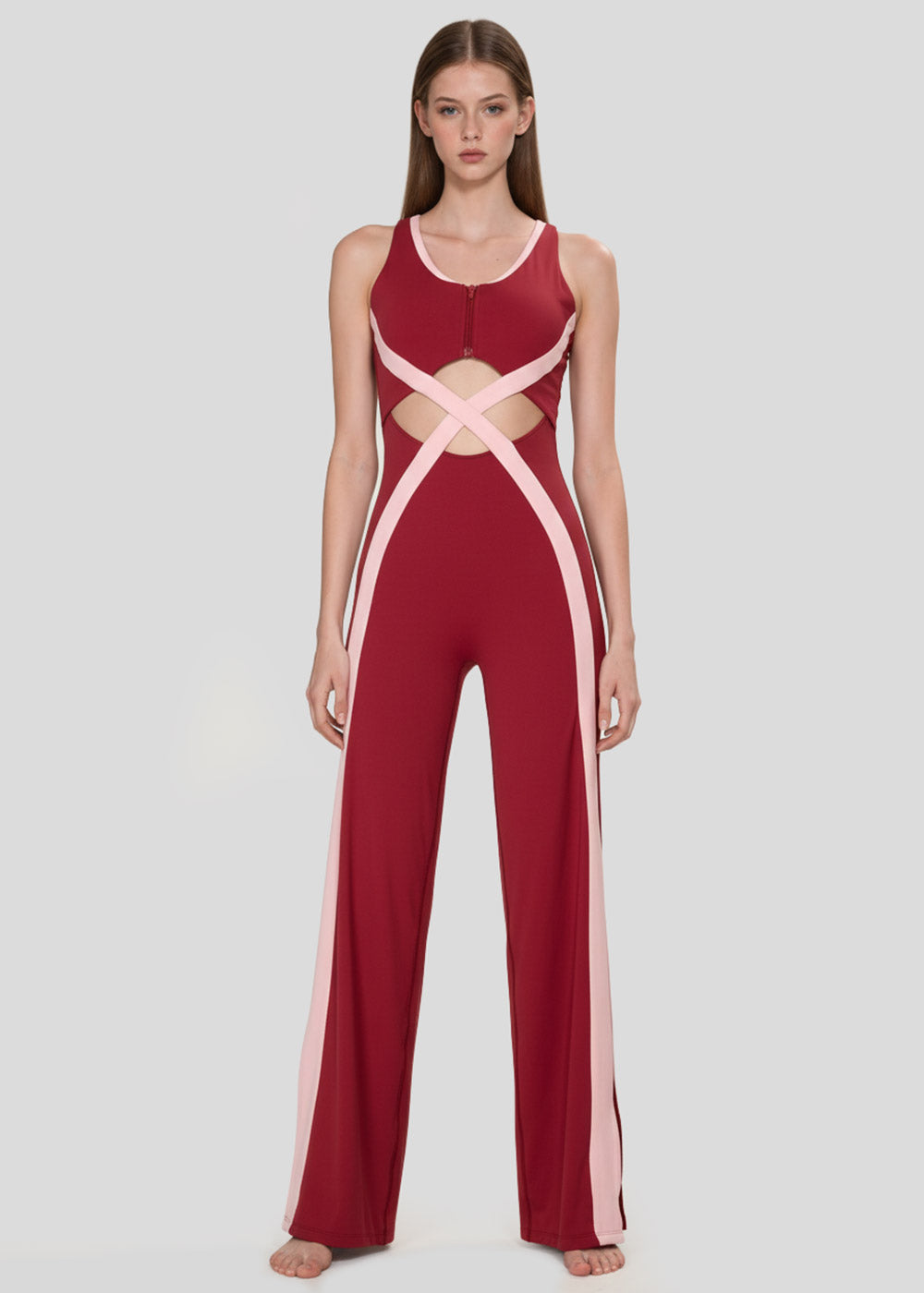 Front view of a model wearing a burgundy and blush pink colorblock wide-leg jumpsuit with waist cutouts, crossover detailing, and a front zipper