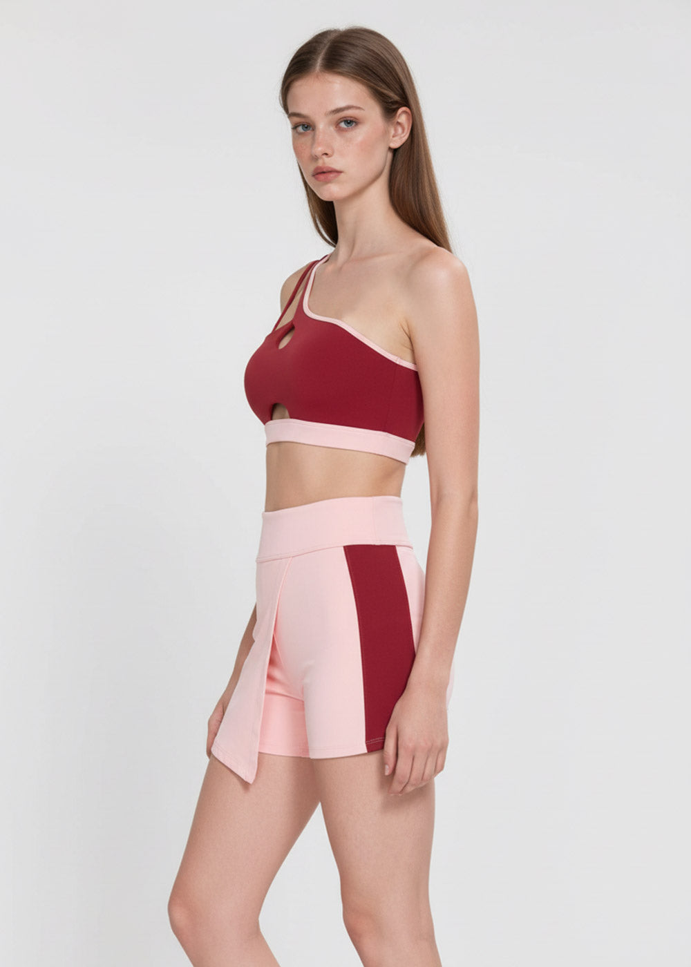 Women's burgundy one-shoulder sports bra with cutout details paired with high-waisted blush pink active shorts with contrast side panels