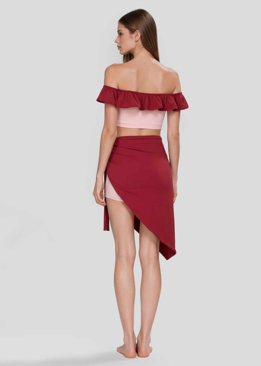 Back view of women's blush pink off-shoulder ruffle crop top with burgundy trim, pink fitted shorts and burgundy asymmetrical wrap skirt